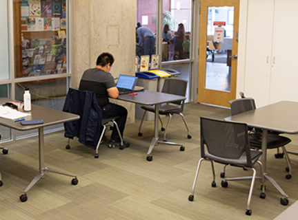 The Mount Pleasant meeting room looking toward the glass doors leading to the main library area. There are four small tables with one or two chairs at them there are two of the tables have a person seated at them, faced away from the camera.