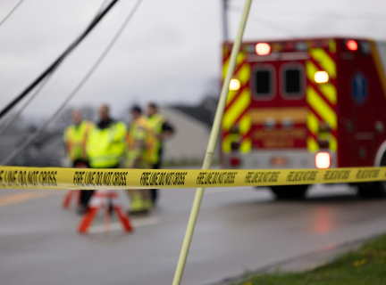 Yellow caution tape reading "Fire Line Do Not Cross" is in focus against a blurred background of emergency personnel gathering next to an ambulance.