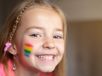 A child smiles at the camera. The child has long blonde hair and is wearing rainbow facepaint and a pink shirt.