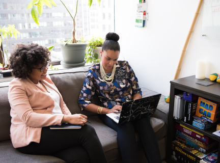 two women sitting on a couch and looking at a laptop screen 