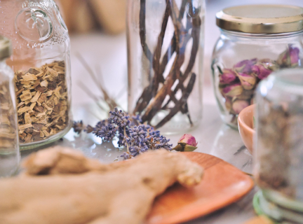 an array of glass jars holding dried herbs and flowers, and a piece of ginger on a wooden plate on a marble countertop