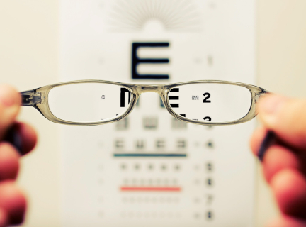 a pair of glasses is being held in front of the camera as if the viewer is seeing through them. an eye examination chart is visible in the distance, with the chart appearing in-focus when viewed through the glasses lenses.