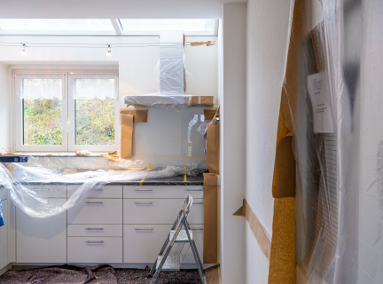 a kitchen space that is clearly undergoing renovation. The walls are white and bare, and the countertops and appliances are wrapped in protective plastic.