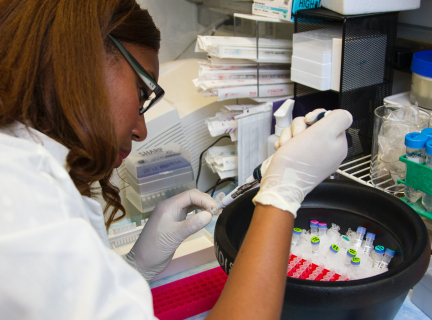 a female microbiologist in a laboratory uses a pipette to transfer DNA samples into a test tube. there is a black round container filled with a series of test tubes on the table in front of her.