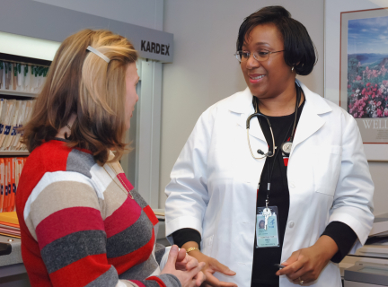 a female healthcare worker in a white lab coat and wearing a stethoscope around her neck explains something to a patient who is wearing a red sweater.