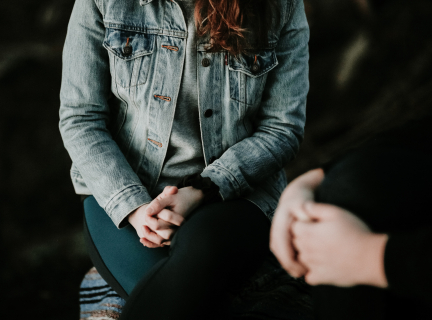 woman in a jean jacket sitting facing another person, both have folded hands on their laps