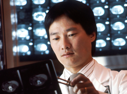 a male radiologist technician handles an x-ray, while standing in front of an array of x-rays on a wall