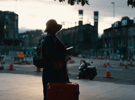 woman wearing coat standing on road with travel luggage during daytime