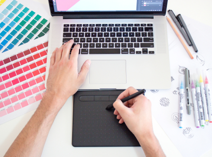 a graphic designer using a laptop and a trackpad to draw, with markers, paper drawings, and colour swatches laid out on the table