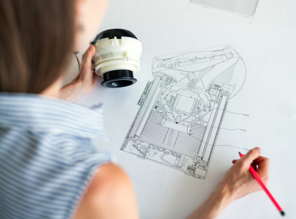 a woman seated before a schematic laid out on a table, while holding a pencil in one hand and the device that the schematic is based off of in the other