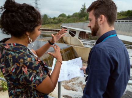 two people standing in front of a waterway looking at technical drawings