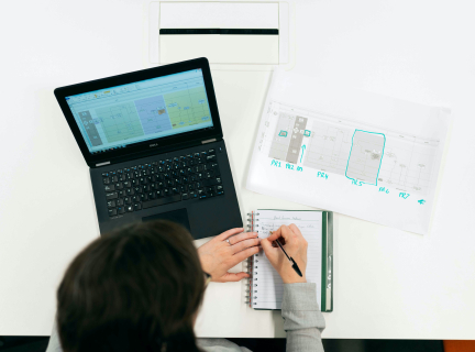 a woman seated at a desk with a laptop displaying a schematic, a paper schematic, and a notebook arrayed in front of her. she is writing in the notebook.