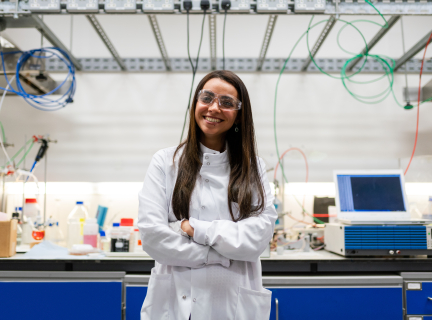 a woman in a white lab coat and goggles stands smiling with crossed arms in front of a laboratory workbench