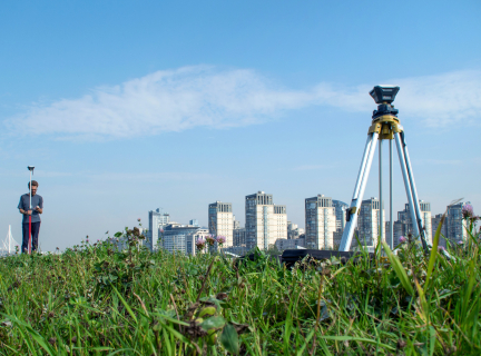 a man holding land surveying gear stands in a field, with a city skyline in the distance.