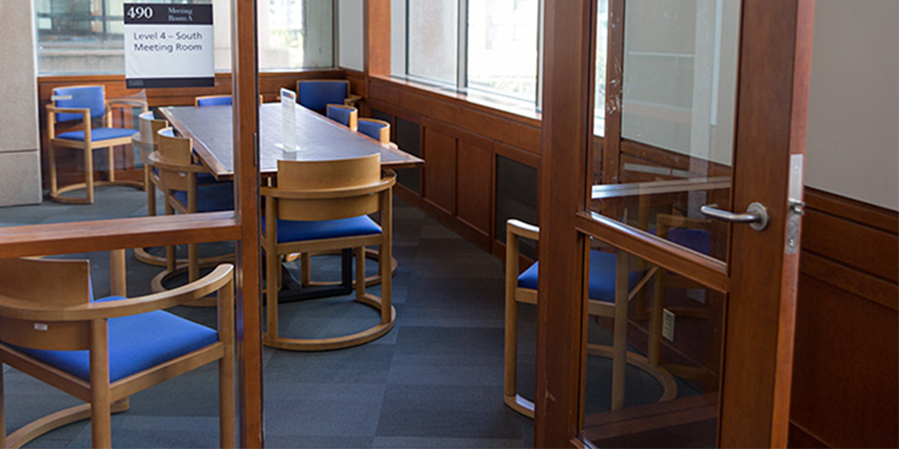 The Central Library L4 South meeting room looking through the door. There is one rectangular table surrounded by six chairs and an additional chair in each of the corners facing inward.