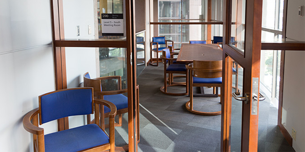 The Central Library L5 South meeting room looking through the door. There is one rectangular table surrounded by six chairs and an additional chair in three of the corners facing inward. A fourth chair is outside the room to the left of the door, facing the camera.