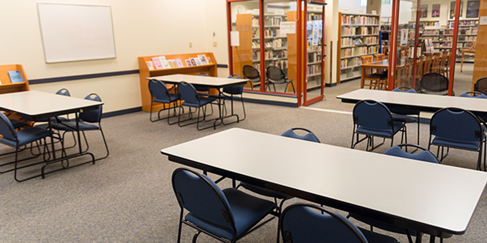 The Champlain Heights meeting room looking toward the glass doors leading to the main library area. There are three small tables in view, each with four chairs, and a small pamphlet shelf and whiteboard on the back wall.