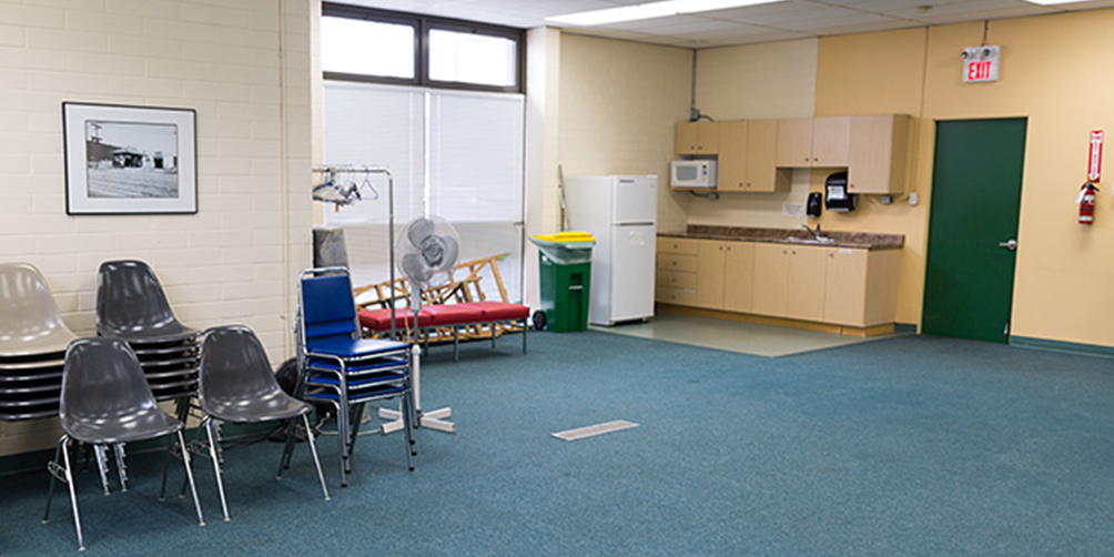 The Fraserview meeting room looking toward the kitchenette in the back corner. There are a number of chairs stacked back wall, along with a few other bits of equipment (coat rack, fan, ladder).