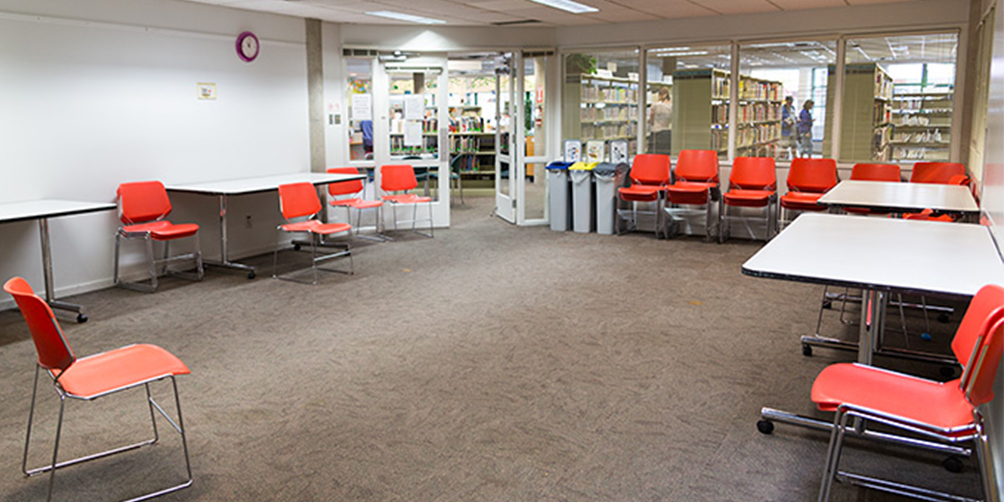 The Hastings meeting room looking toward the glass doors leading to the main library area. There are four small tables around the edge of the room, along with a number of chairs against the wall, some in small stacks.