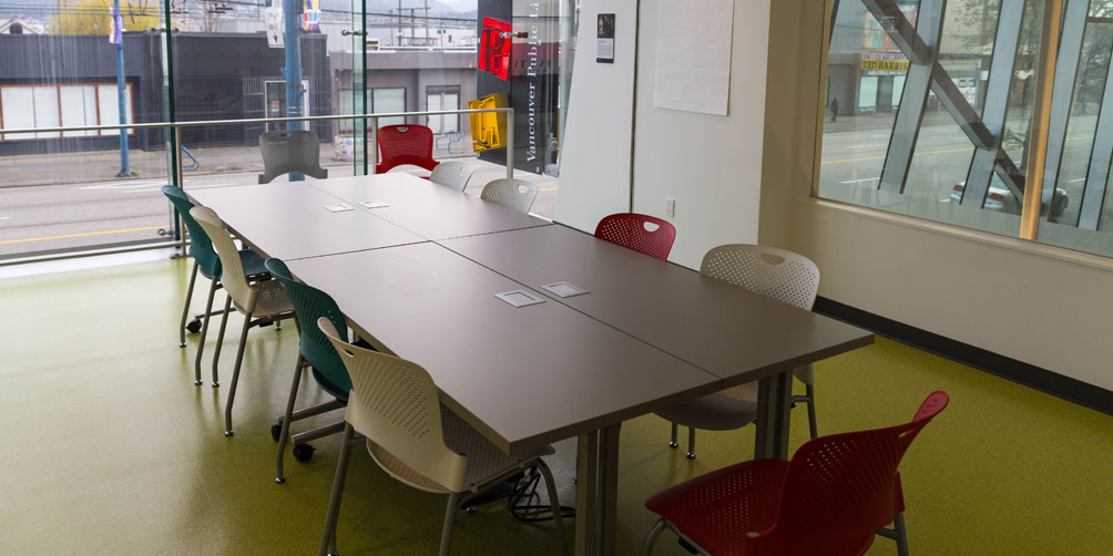 The Nellie Yip Quong Room, looking toward the back glass wall overlooking the street. There is a large rectangular table made up of four smaller tables pushed together. They are surrounded by eleven chairs.