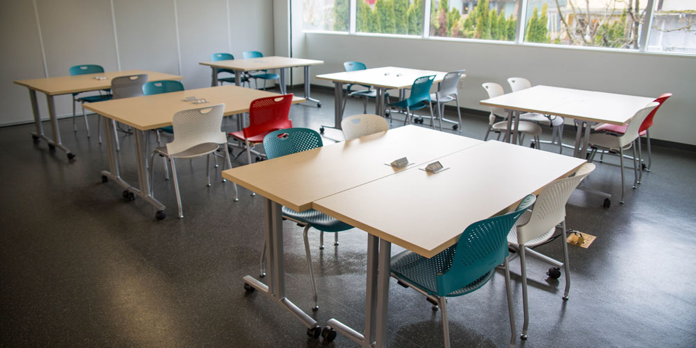 The Wo Soon (Mary) Lee Chan Room looking toward the windowed back wall. There are six square tables, each made of two smaller rectangular tables pushed together. Each table has from 2 to 4 chairs at it.