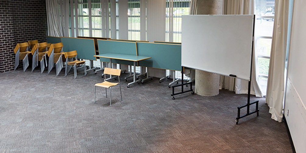 The Renfrew meeting room looking toward the windowed back wall. There is a whiteboard, a small table, and a chair. There are several stacks of chairs and folded tables against the wall below the windows.