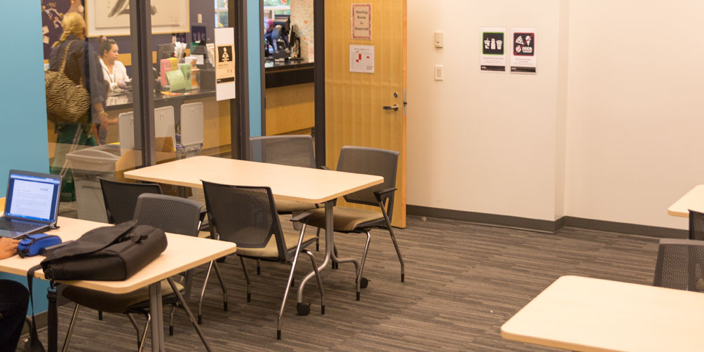 The Terry Salman meeting room looking toward the doors leading to the main library area. There are portions of four small tables in view, each with up to four chairs, with a laptop and laptop case on one of them. Through the glass window next to the door you can see a library worker at a desk and a patron talking with them, facing away from the camera.