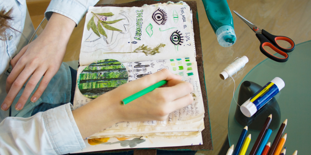 An image of a hand drawing in a sketchbook on a table with other art supplies.