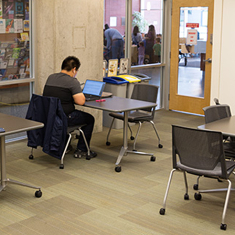 The Mount Pleasant meeting room looking toward the glass doors leading to the main library area. There are four small tables with one or two chairs at them there are two of the tables have a person seated at them, faced away from the camera.